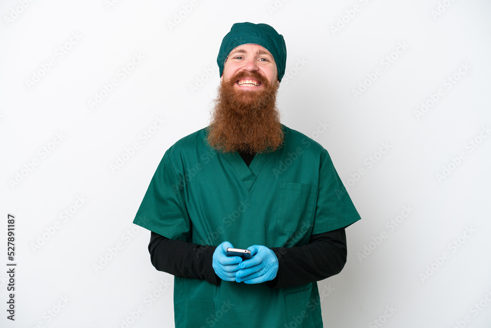 Surgeon redhead man in green uniform isolated on white background sending a message with the mobile Surgeon redhead man in green uniform isolated on white background sending a message with the mobile