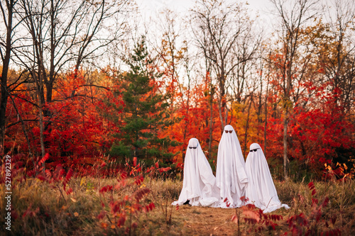Three children dressed as ghosts wearing sunglasses play in front of colorful autumn foliage
