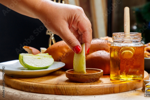 A woman's hand dips a piece of apple in honey in honor of the celebration of Rosh Hashanah near honey and challah and traditional food. photo