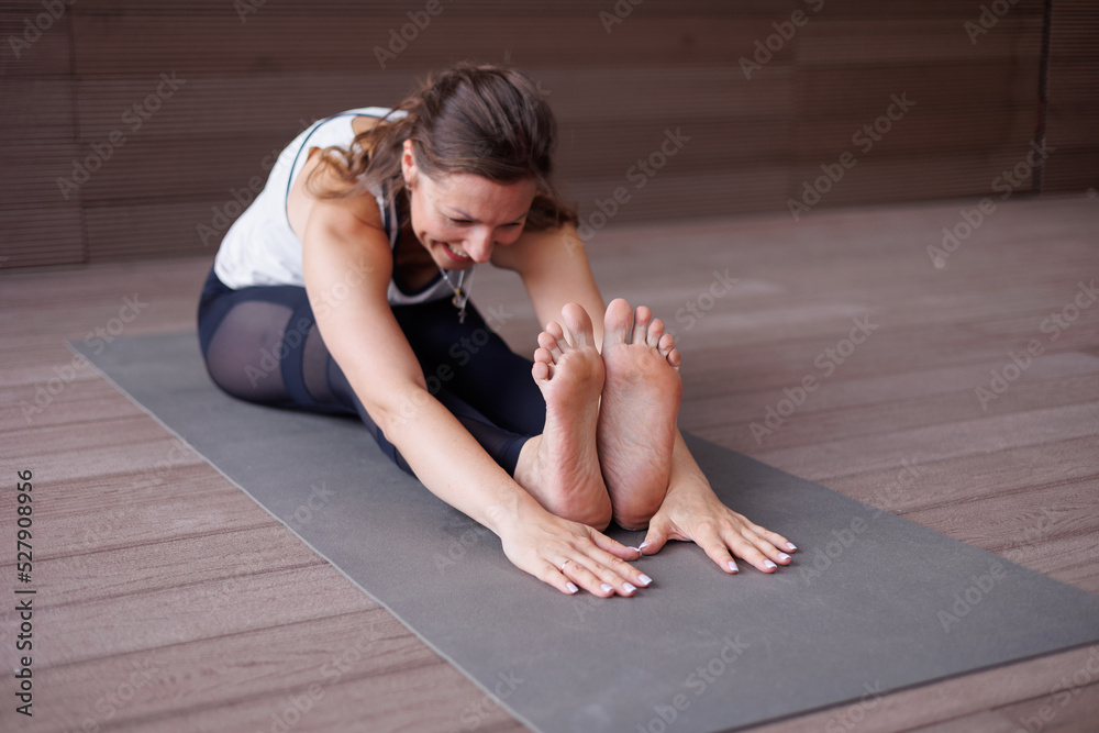 Obraz premium Beautiful strong muscular woman doing stretching on mat on terrace outdoors. Healthy young adult girl in sportswear stretch alone. Fit thin gimnast doing yoga, training on mat