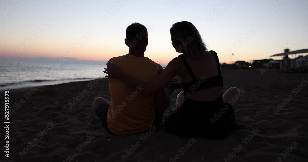 Husband and wife are sitting on beach daughter runs up to and hugs them at sunset Husband and wife are sitting on beach daughter runs up to and hugs them at sunset