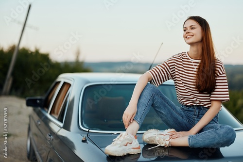 A young woman travels alone in her car on the roads in the countryside and relaxes sitting on the hood watching the sunset © SHOTPRIME STUDIO