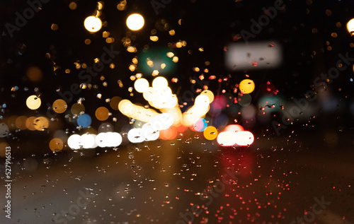 Blurred lights of cars at night behind the windshield with water drops. Background with lights beyond the depth of field.
