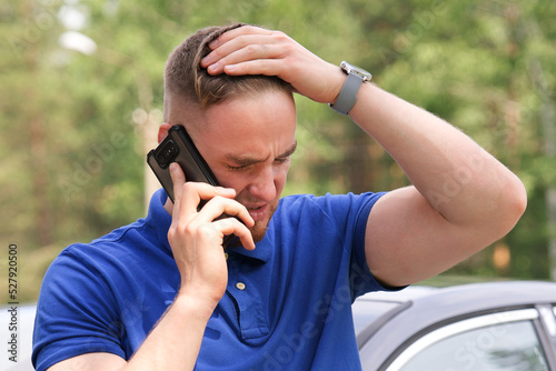 Sad upset guy driver is standing near broken car after road accident, crash, calling emergency on cell mobile phone talking on smartphone. Young desperate man got into accident holding head with hand