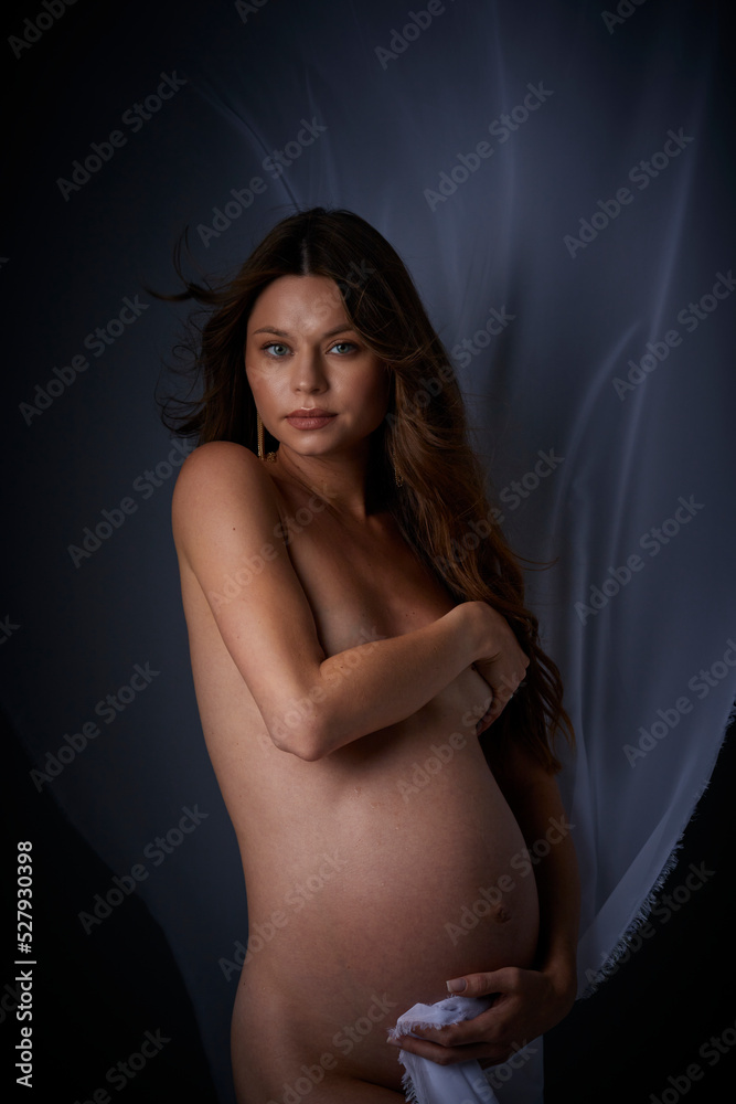 Naklejka premium Beautiful young pregnant serious sexy woman with long curly hair and big belly, without clothes sitting in dark studio and covers her breasts with her hand
