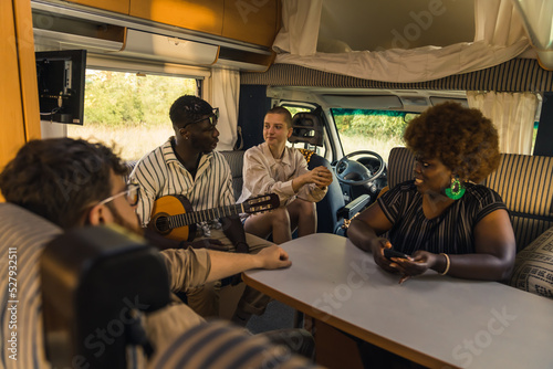 Active interracial group of four good friends on a summer road traveling, sitting inside their comfortable modern motorhome, playing the guitar, chatting and laughing. RVs interior. High quality photo