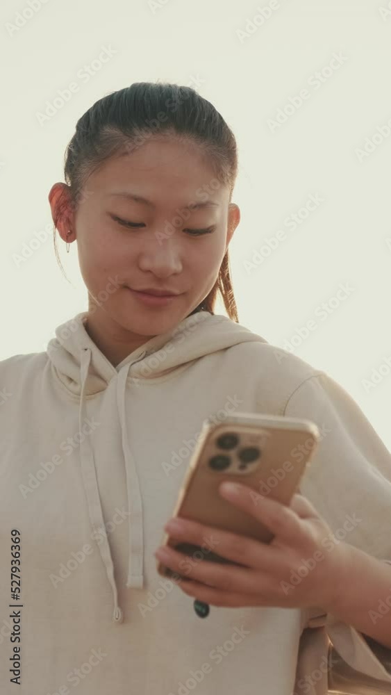 VERTICAL VIDEO: Asian girl in sportswear uses the phone while standing on the waterfront at morning time VERTICAL VIDEO: Asian girl in sportswear uses the phone while standing on the waterfront at morning time