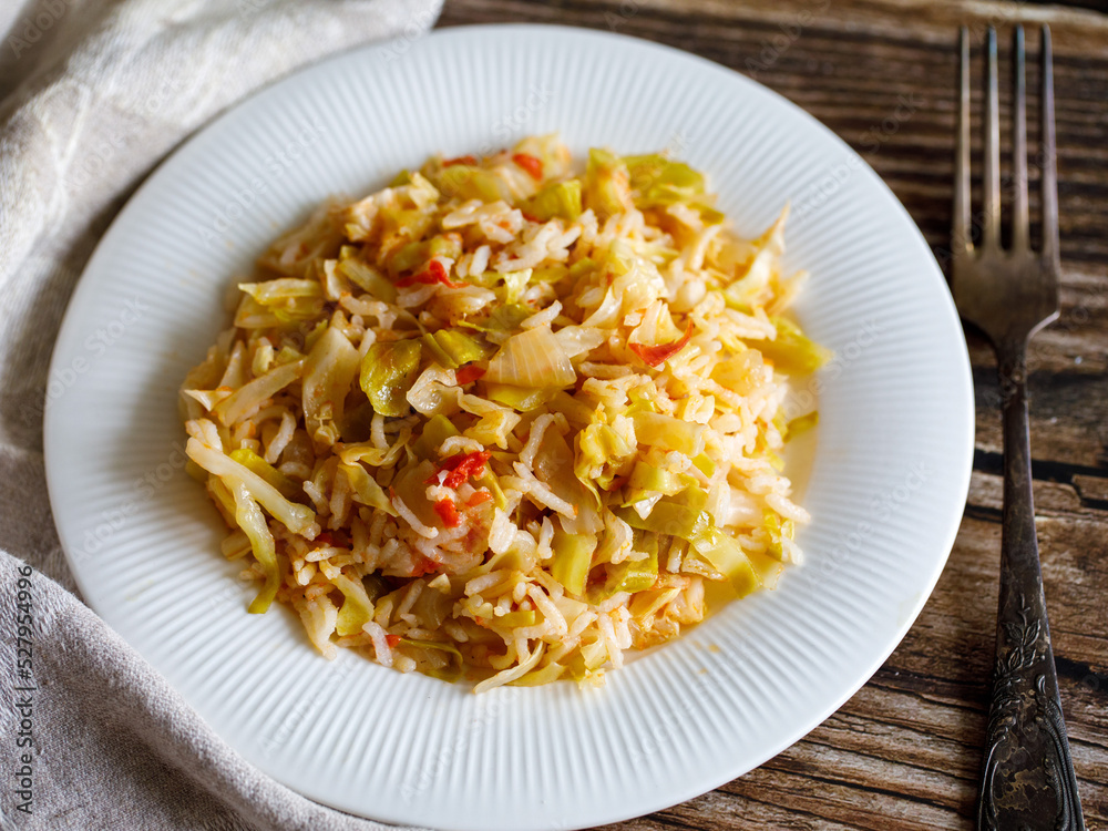 Traditional homemade Mediterranean Crete cuisine rice with vegetables cabbage, onion and tomato on white plate with fork and napkin on old wooden table. Vegetarian healthy lunch, dinner idea. Top view Traditional homemade Mediterranean Crete cuisine rice with vegetables cabbage, onion and tomato on white plate with fork and napkin on old wooden table. Vegetarian healthy lunch, dinner idea. Top view