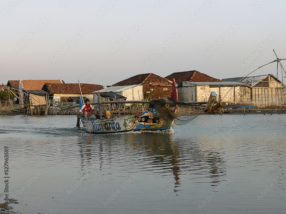 Jepara 09-02-2022: Photo of a fishing boat speeding up the river, driven by a man, with several residential houses in the background