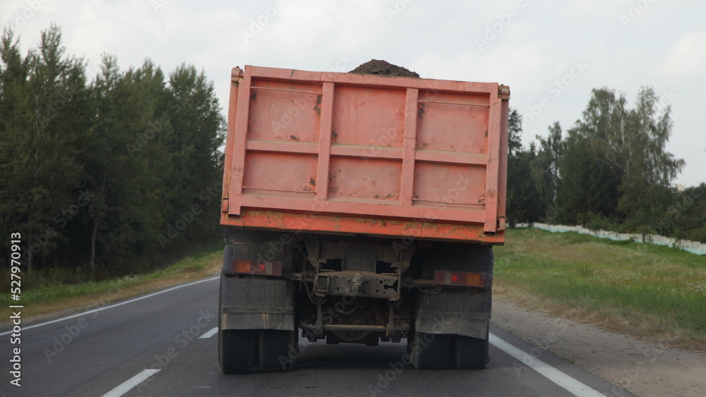 Heavy tilted overloaded dump truck carries ground on a European countryside asphalt road on green trees background at summer day