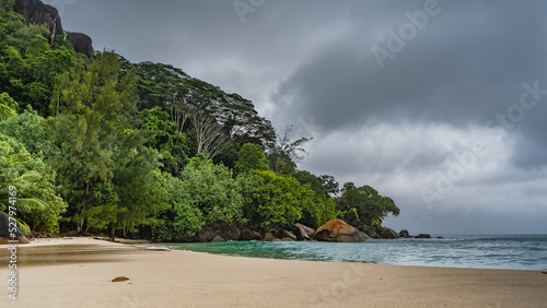 Rainy foggy day on a tropical island. Boulders in the turquoise ocean, clean wet sand of the beach. Lush green vegetation on the hill. Gray clouds in the sky. Seychelles. Mahe