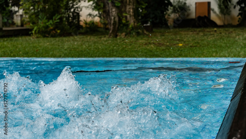 A fragment of an outdoor Jacuzzi. The blue water is bubbling  splashes are flying in the air. The soft background is a green lawn.