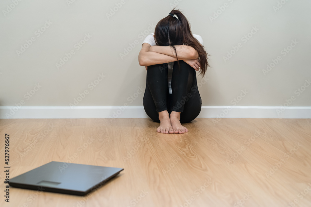 Fototapeta premium Depressed and stress, sad asian young woman,girl cover face with hand, sitting on floor, weighing on scale in front of her. Gain lose weight and overweight fat. People diet, unhealthy, unhappy concept