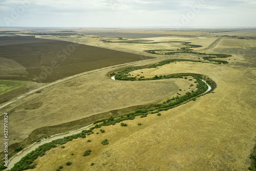 a winding muddy river with overgrown green banks in a sun-scorched steppe rural landscape