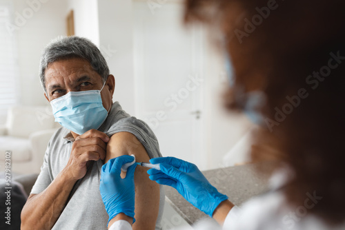 Mixed race female doctor wearing mask giving vaccination to senior man at home photo