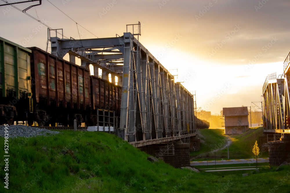 A railway bridge on which a train with wagons moves against the background of a bright sun