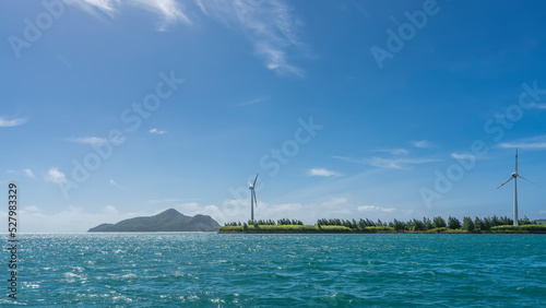 There are wind turbines on an island in the ocean. Turquoise water sparkles in the sun. Light clouds in the blue sky. Copy space. Seychelles