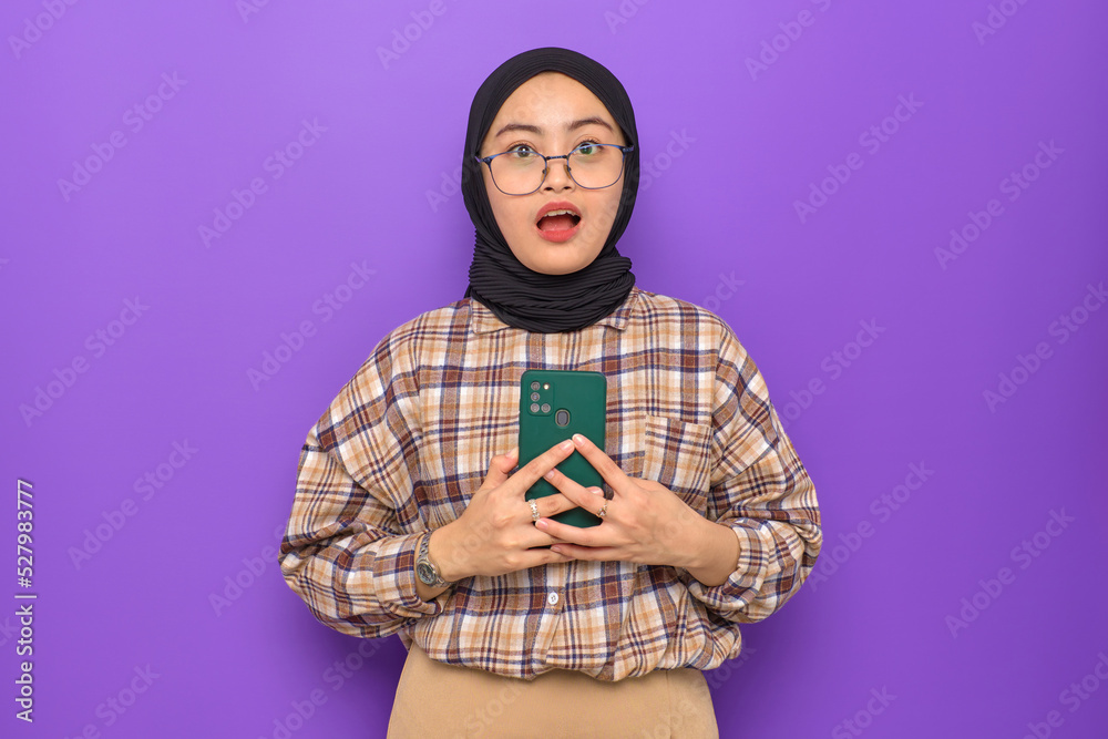 Shocked young Asian woman in plaid shirt holding a mobile phone, looking at camera isolated on purple background Shocked young Asian woman in plaid shirt holding a mobile phone, looking at camera isolated on purple background