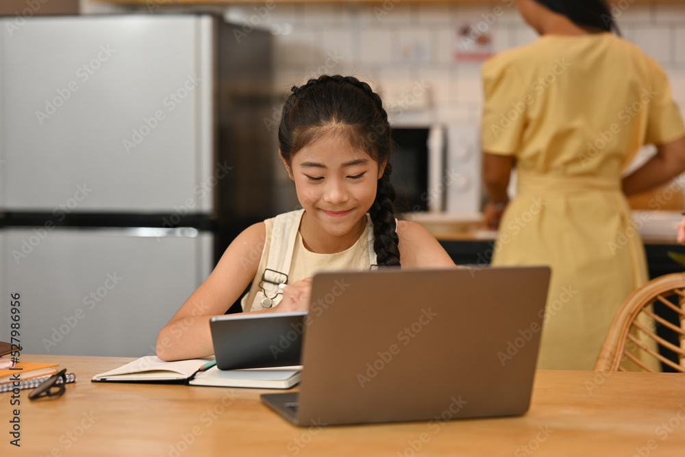 Happy Asian family, Mom is busy cooking at the kitchen while her daughter study with laptop and tablet, E-learning, Online education concept. Happy Asian family, Mom is busy cooking at the kitchen while her daughter study with laptop and tablet, E-learning, Online education concept.