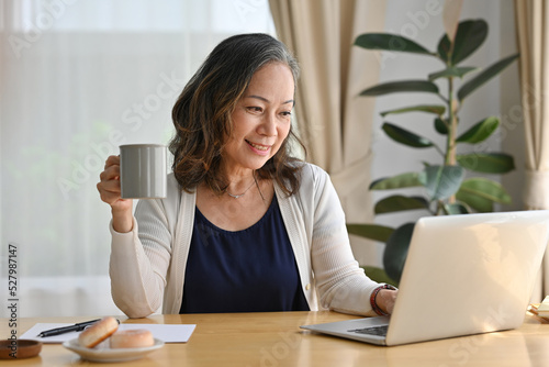 Happy elderly lady using laptop for webinar while drinking coffee, having working online on Internet, copy space, Work at home concept. © saltdium