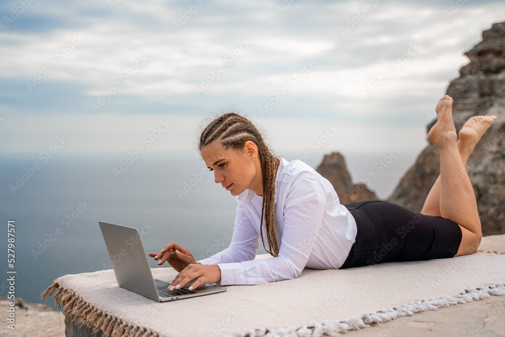 A woman is lying and typing on a laptop keyboard on a terrace with a beautiful sea view. Wearing a white blouse and black skirt. Freelance travel and vacation concept, digital nomad.