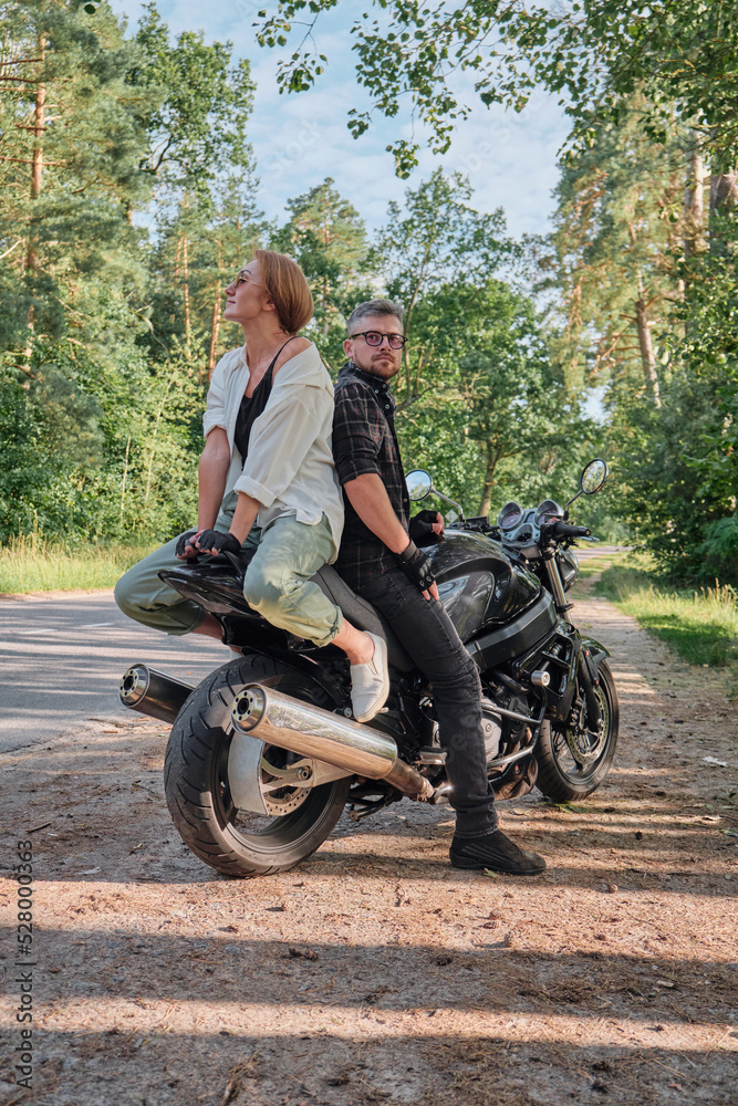 Naklejka premium Middle age couple talking and having fun, sitting on a motorcycle, traveling together on a forest road