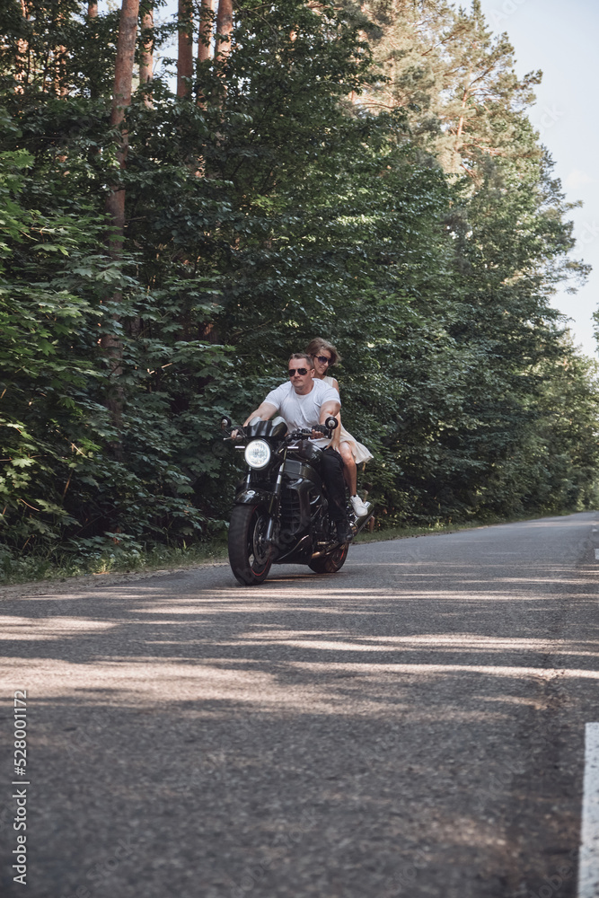 Obraz premium A young happy couple rides a motorcycle on an asphalt road in the forest, freedom and speed