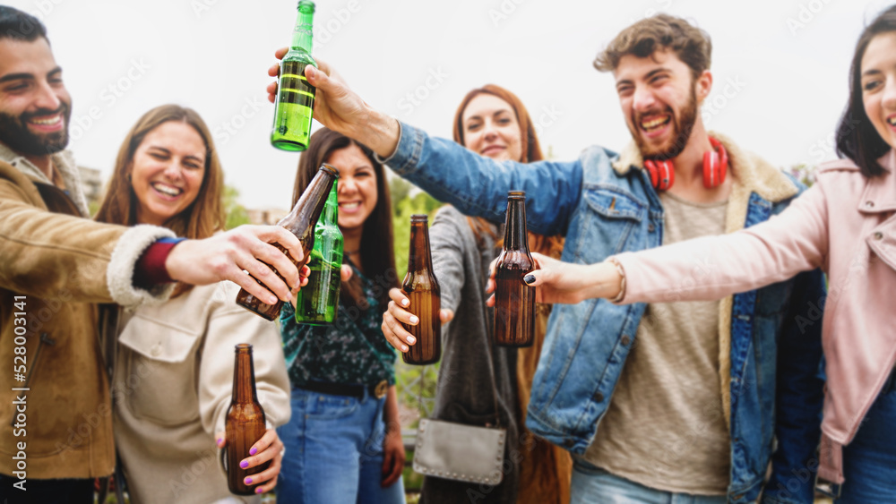 Gathering of millennial generation people having fun outdoors celebrating friendship toasting with beers - group of young people raising beers in the sky - food and drink lifestyle concept