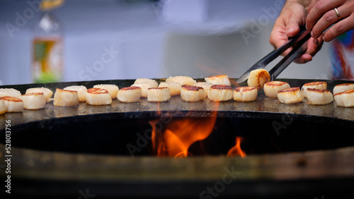 Delicious scallops being flipped over grilling on round steel iron firepit hearth table surface brazier barbecue, shot with shallow dept of field