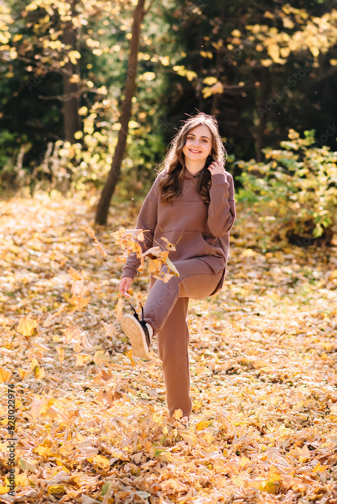 Young woman in hoodie sweater kicks up leaves in autumn park. Sunny weather. Fall season. Young woman in hoodie sweater kicks up leaves in autumn park. Sunny weather. Fall season.