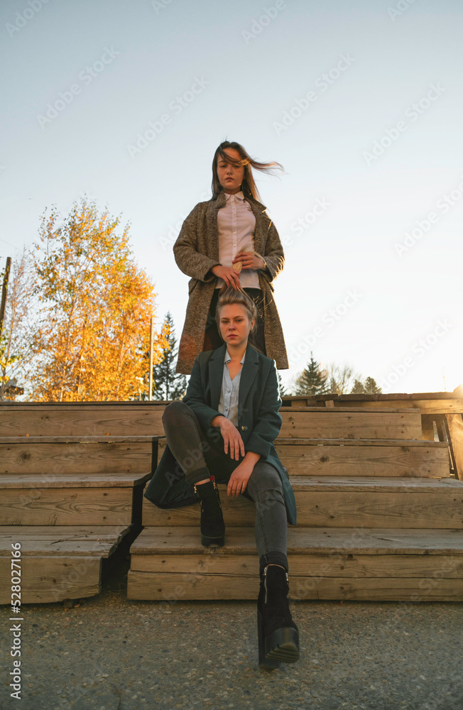 Two friends are walking outdoors in autumn, girls sitting at a wooden fence. One of them is holding the other's hair in her hands Two friends are walking outdoors in autumn, girls sitting at a wooden fence. One of them is holding the other's hair in her hands