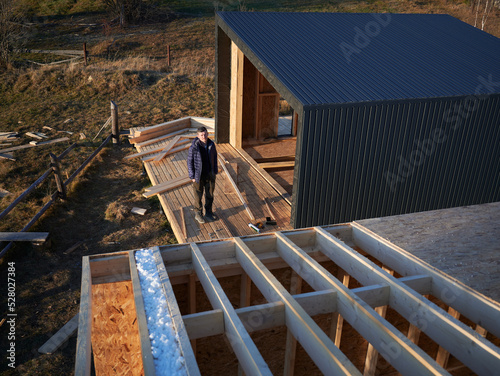 Male architect building wooden frame house in the Scandinavian style barnhouse. Man standing on terrace at construction site, inspecting quality of work in the evening.