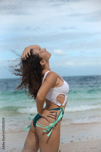 Portrait of a beautiful female in a bikini on a tropical beach. A brunette girl in swimsuit enjoy at the sea. A curly woman tourist posing on a tropical beach and enjoys during her summer holiday