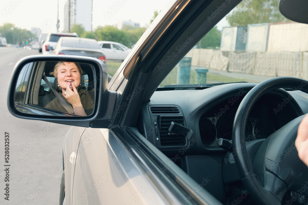 Middle aged woman with short hair driving a car paints her lips with lipstick. The woman driving making make-up. Reflection of a woman in the rearview mirror.