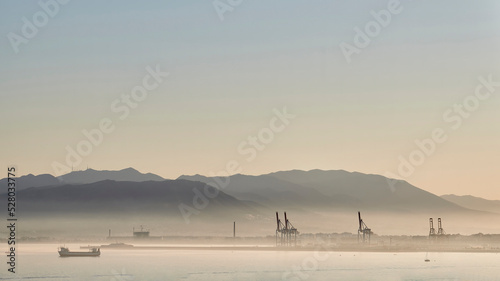Sunset in the port of Malaga with fog caused by humidity with cranes, ships sailing and mountains behind © MSCT