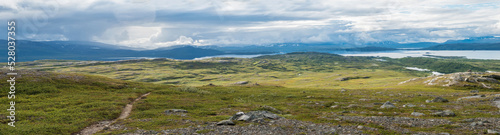 Panoramic landscape with beautiful Virihaure lake, snow capped mountain, birch trees and footpath of padjelantaleden hiking trail. Sweden, Lapland wild nature. Summer cloudy day