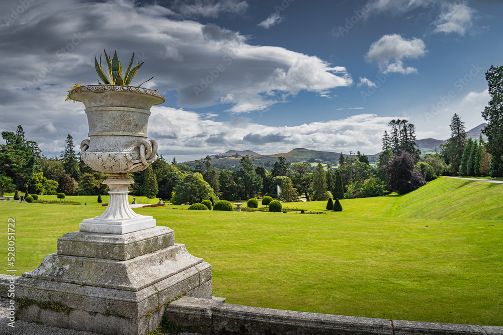 An ornate pot with flowers, wide green lawn with bushes in Powerscourt gardens, forest and Sugerloaf mountain in the background, Wicklow, Ireland