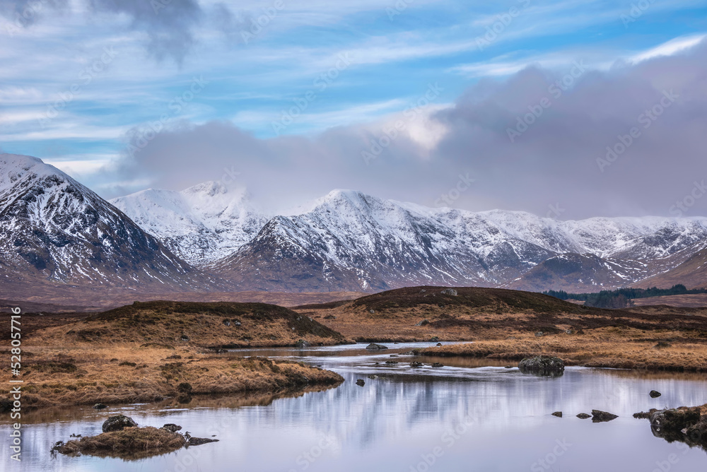 Majestic Winter panorama landscape image of mountain range and peaks viewed from Loch Ba in Scottish Highlands with dramatic clouds overhead