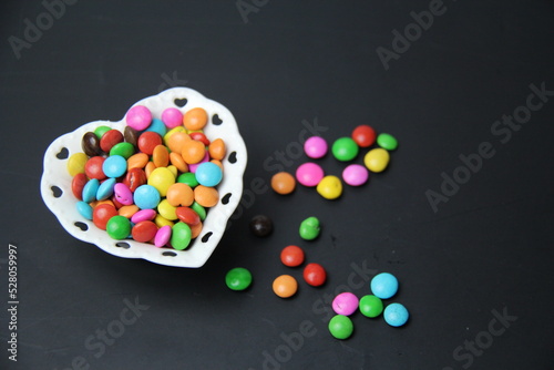 Colorful sugar coated chocolate candies in a bowl, white with heart shape on a black background with space for text, selective focus, top view.