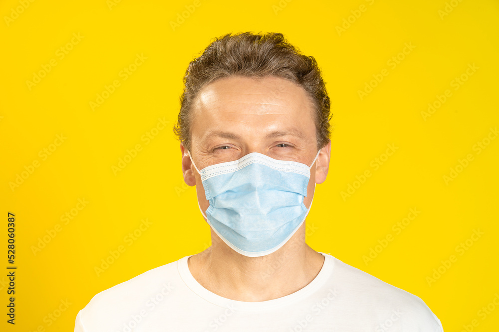 Happy, excited young man wearing a medical mask on his face. Sick young man in face mask wearing white t-shirt isolated on yellow background. A smiling caucasian man in protective face mask Happy, excited young man wearing a medical mask on his face. Sick young man in face mask wearing white t-shirt isolated on yellow background. A smiling caucasian man in protective face mask