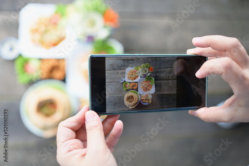 A woman holding a smartphone or mobile phone takes pictures of a set Thai dishes and shares it on social networks before eating. Food served on wooden table consists of vermicelli salad,steamed,etc. 