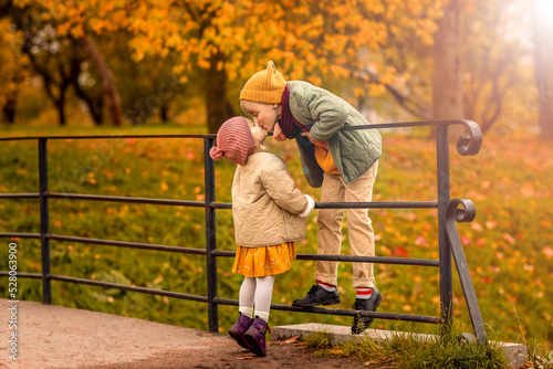 Autumn friendly romantic kiss cute couple children boy and girl on bridge in park. Kids wearing casual stylish clothes. Full body view.