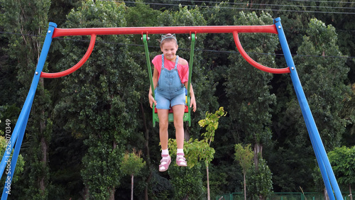 smiling, happy eight year old girl swinging on a swing, outdoors, in a park, summer, hot day during a vacation. Little girl having fun on a swing outdoor. High quality photo