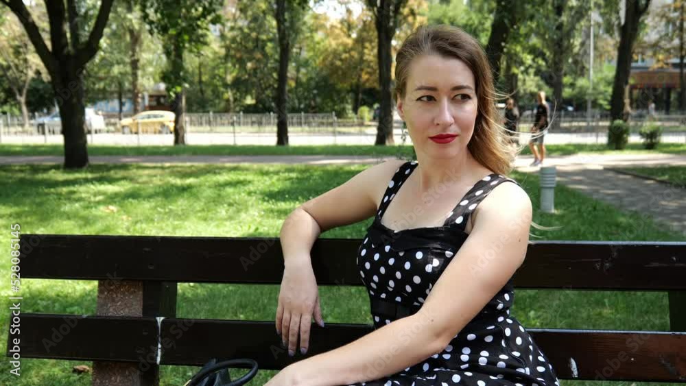 A young woman in a black and white dress sits on a bench in a city park. Slow motion portrait woman relaxing sitting A young woman in a black and white dress sits on a bench in a city park. Slow motion portrait woman relaxing sitting