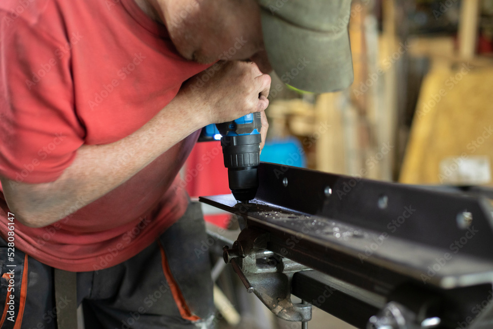 A man drills holes in the metal. The guy works in the garage. A man drills holes in the metal. The guy works in the garage.