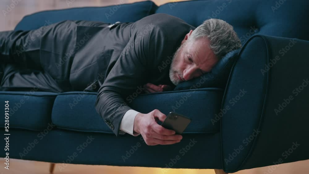 Tired businessman lying on sofa, reading news about company bankruptcy on phone Tired businessman lying on sofa, reading news about company bankruptcy on phone