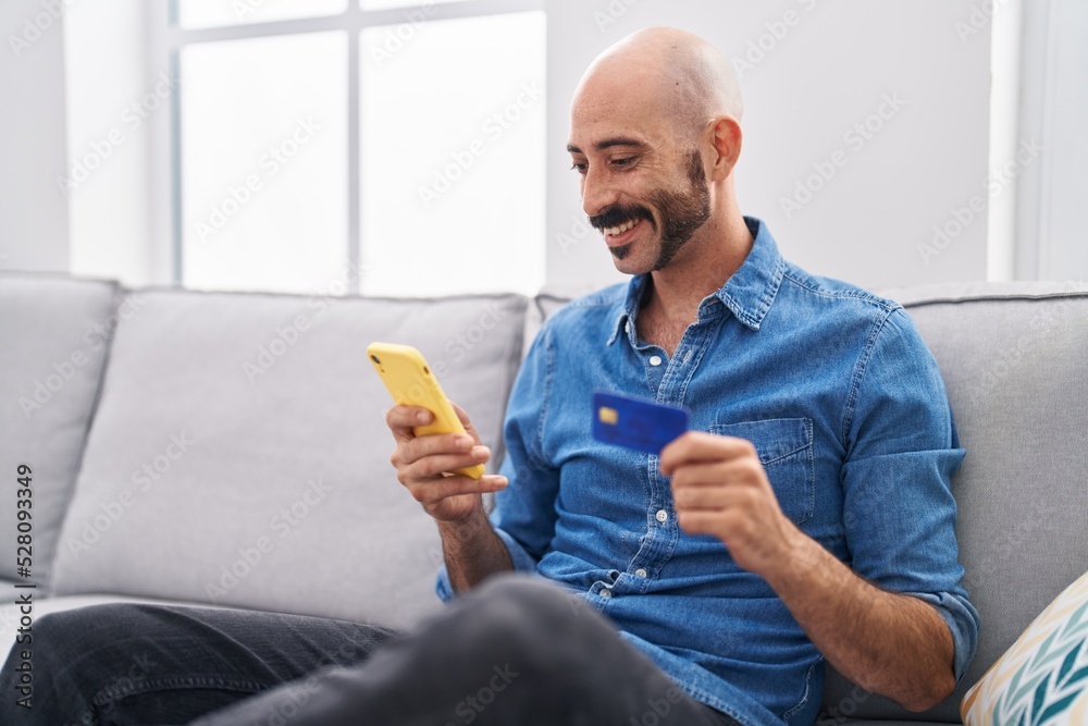 Young hispanic man using smartphone and credit card sitting on sofa at home Young hispanic man using smartphone and credit card sitting on sofa at home