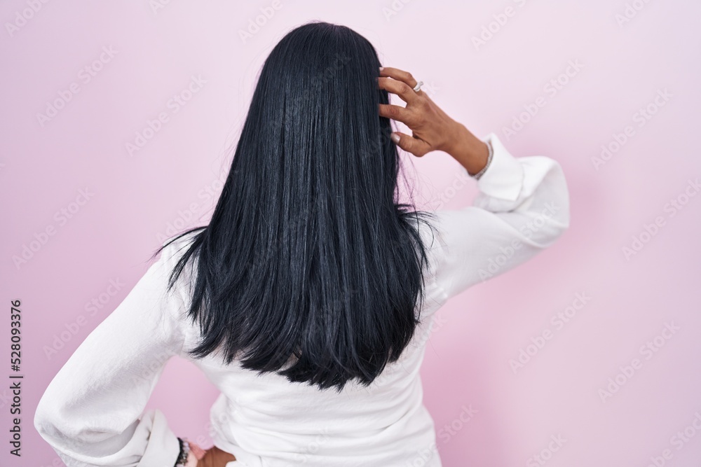 Mature hispanic woman standing over pink background backwards thinking about doubt with hand on head Mature hispanic woman standing over pink background backwards thinking about doubt with hand on head