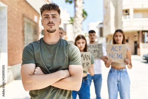 Young hispanic activist man with arms crossed gesture standing with a group of protesters holding banner protesting at the city Fototapet