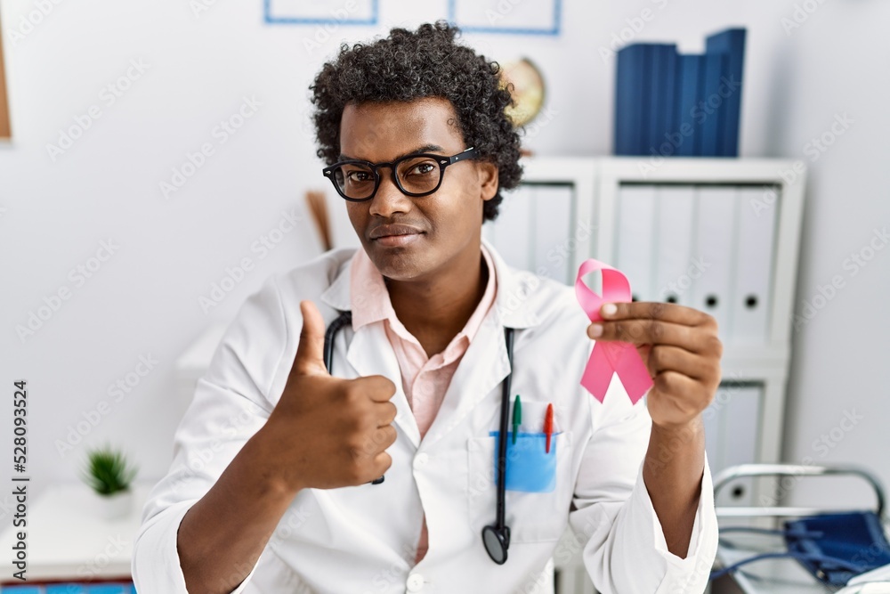 African doctor man holding pink cancer ribbon smiling happy and positive, thumb up doing excellent and approval sign African doctor man holding pink cancer ribbon smiling happy and positive, thumb up doing excellent and approval sign